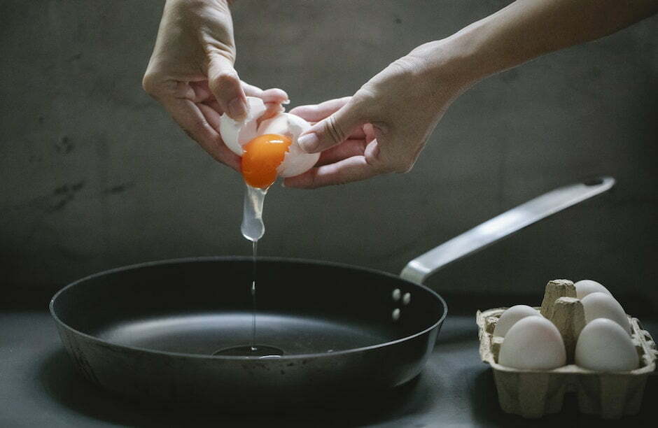 Crop anonymous female breaking egg in pan placed on table while cooking breakfast